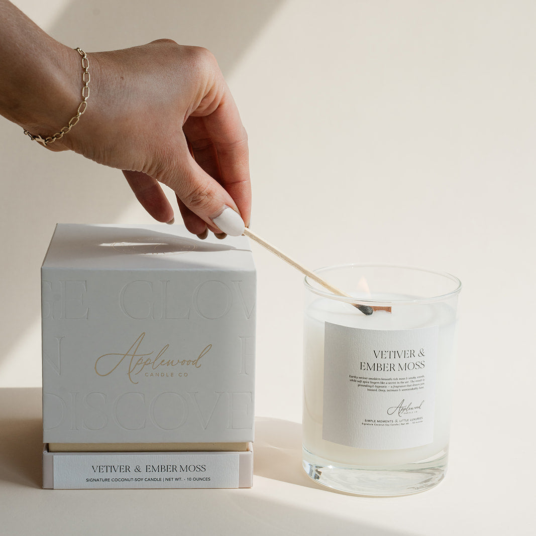 Vetiver & Ember Moss candle in clear glass jar being lit with box on beige background.