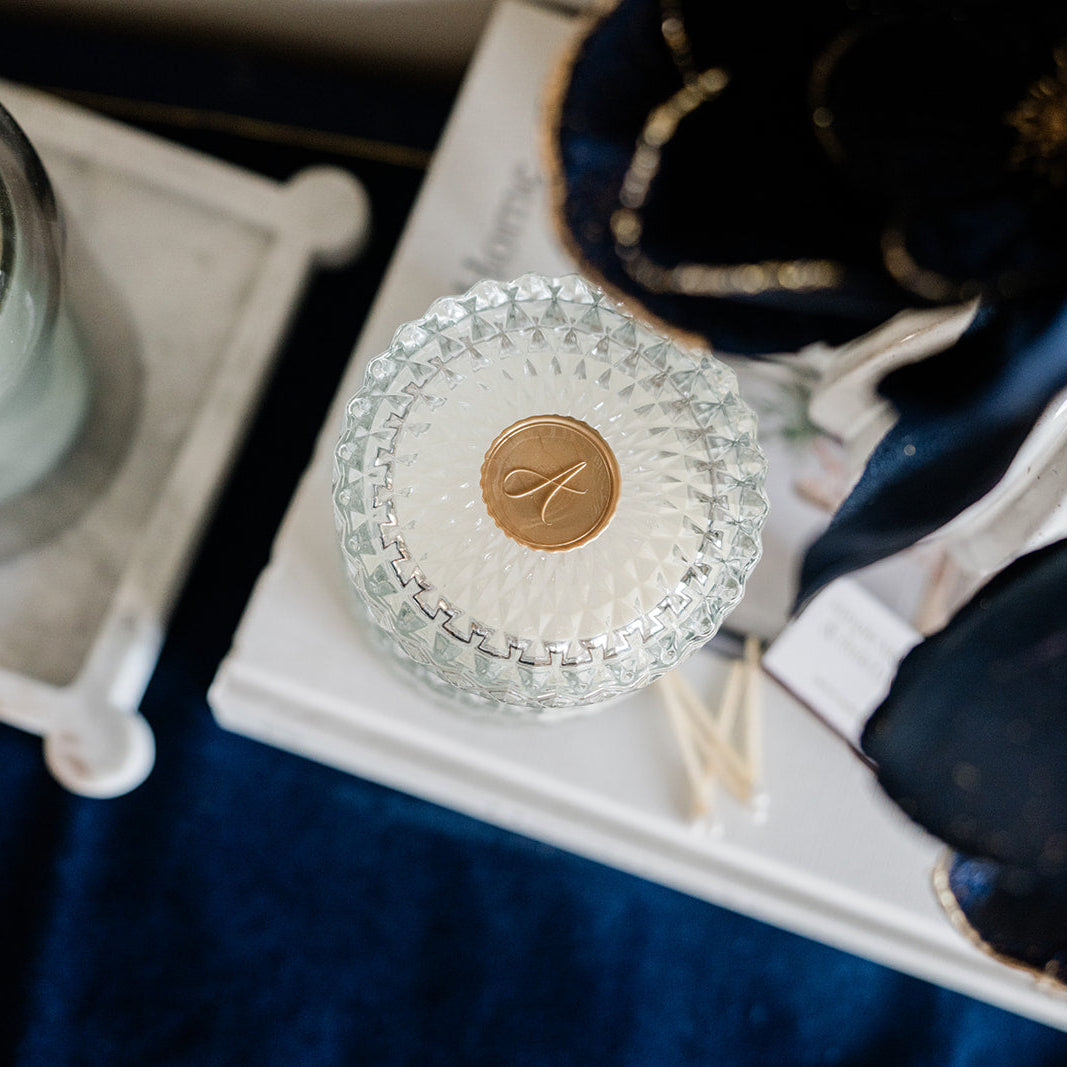 Decorative glass item with a gold emblem on a white surface, with a candle and dark fabric in the background.