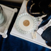 Decorative glass item with a gold emblem on a white surface, with a candle and dark fabric in the background.