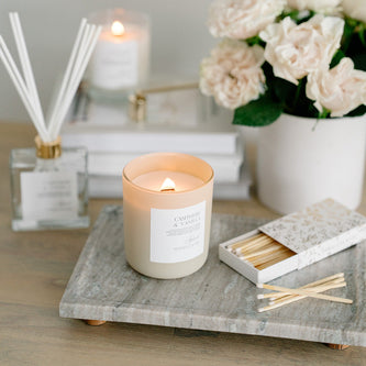 Lit Cashmere & Vanilla candle in frosted glass jar with matches beside it, reed diffuser, signature candle, and candle snuffer in background.