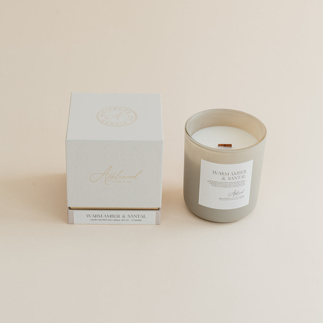 Warm Amber & Santal candle in frosted glass jar with box on beige background.