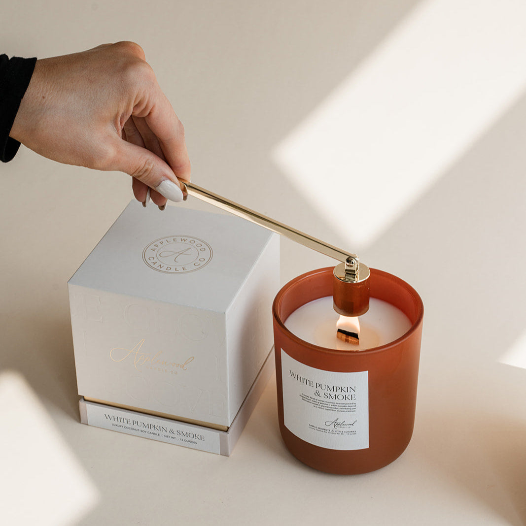 White Pumpkin &amp; Smoke candle in frosted glass jar with box and gold candle snuffer on beige background.