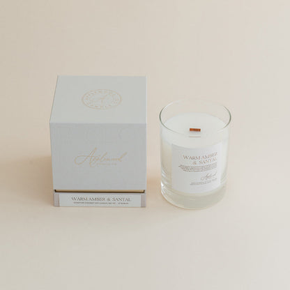 Warm Amber &amp; Santal candle in clear glass jar with box on beige background.