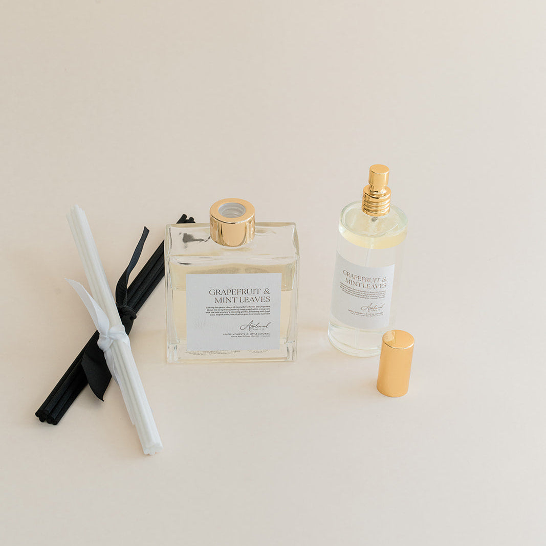 Luxury Flameless reed diffuser and linen spray in glass bottles with gold details, styled on beige backdrop.