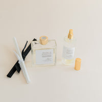 Luxury Flameless reed diffuser and linen spray in glass bottles with gold details, styled on beige background.
