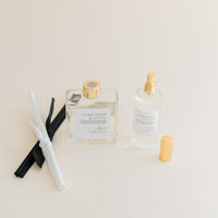 Luxury Flameless reed diffuser and linen spray in glass bottles with gold details, styled on beige backdrop.