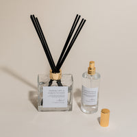 Luxury Flameless reed diffuser and linen spray in glass bottles with gold details, styled on beige backdrop.